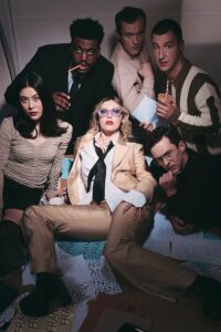 An office scene of 6 people huddled togather with a central female character sprawled out on a table with a seductive look on her face.