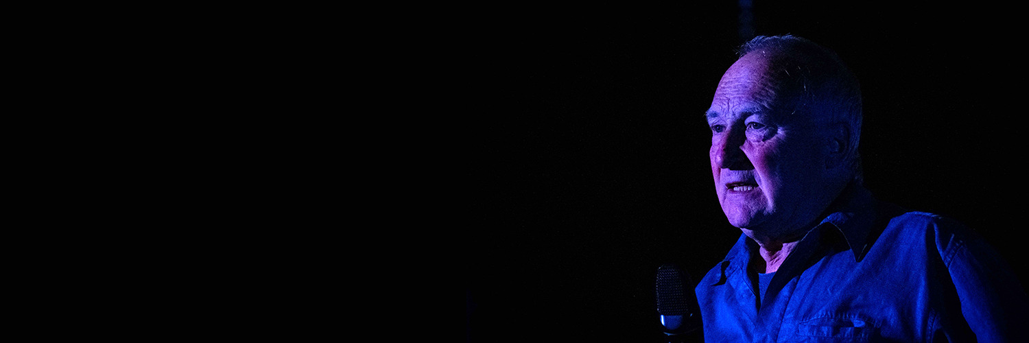 Catalpalyst_Banner A man is shown from the shoulders up, wearing an electric blue shirt against a nearly pitch black background. The subject is illuminated against the darkness by dramatic blue and purple stage lighting. He is looking to the left of the camera with a pensive expression. Deep shadows cover much of his face and clothing, creating a high-contrast, surreal atmosphere. A microphone is visible in the lower left foreground, suggesting a speech or performance.