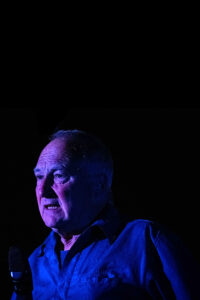 A man is shown from the shoulders up, wearing an electric blue shirt against a nearly pitch black background. The subject is illuminated against the darkness by dramatic blue and purple stage lighting. He is looking to the left of the camera with a pensive expression. Deep shadows cover much of his face and clothing, creating a high-contrast, surreal atmosphere. A microphone is visible in the lower left foreground, suggesting a speech or performance.